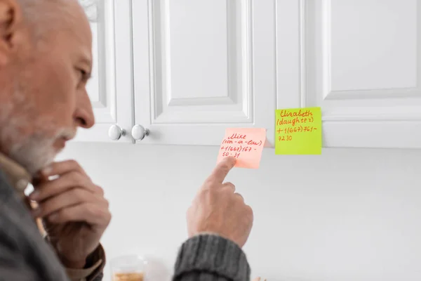 Blurred man suffering from memory loss and pointing at sticky notes with phone numbers and names in kitchen — Stock Photo