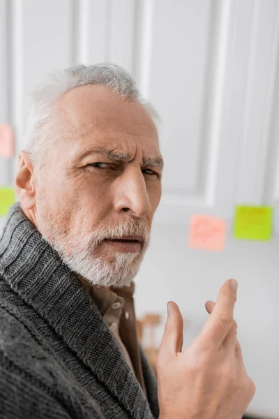 Thoughtful man with alzheimer disease looking at camera near blurred sticky notes in kitchen — Photo de stock