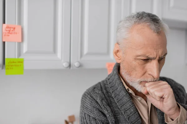 Aged man with alzheimer syndrome holding fist near face while thinking near blurred sticky notes in kitchen — Stock Photo