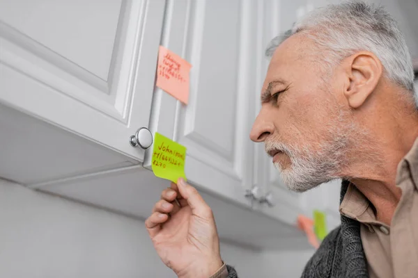 Senior man with alzheimer syndrome looking at sticky note with name and phone number in kitchen — Stock Photo