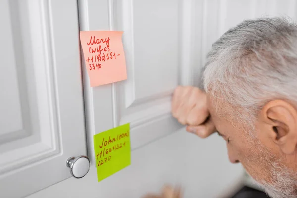 Stressed man suffering from memory loss and standing near sticky notes with names and phone numbers in kitchen — Stock Photo