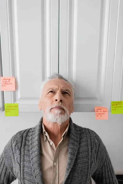 Senior man suffering from memory loss and looking up while standing near sticky notes with names and phone numbers in kitchen — Foto stock