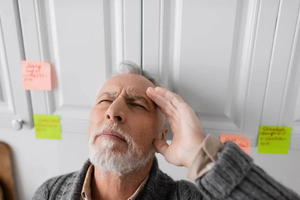 Senior man with closed eyes and hand near head standing near blurred sticky notes while suffering from memory loss — Stock Photo