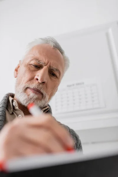 Low angle view of bearded senior man with alzheimer disease writing in blurred notebook at home — Stock Photo
