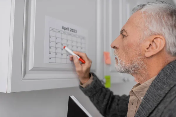 Вид сбоку пожилого человека с альфа-синдромом, указывающего войлочной ручкой на календарь на кухне — стоковое фото