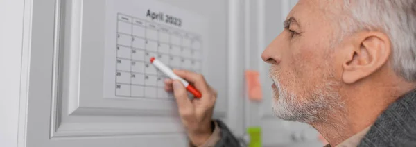 Вид сбоку пожилого человека с альфа-болезнью, указывающего войлочной ручкой на календарь на кухне, баннер — стоковое фото
