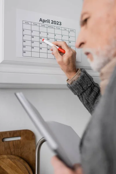 Blurred man with alzheimer syndrome holding blurred notepad and pointing with felt pen at calendar in kitchen — Stock Photo