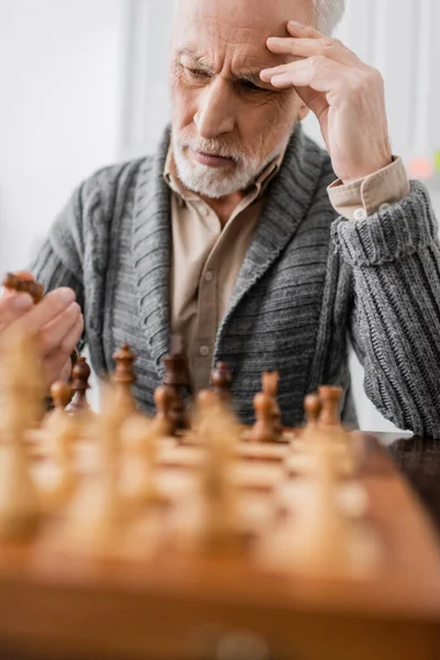 Thoughtful senior man suffering from memory loss and looking at chess figure on blurred foreground — Photo de stock