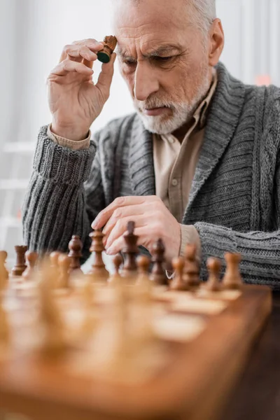Aged man sick on alzheimer syndrome holding figure while thinking near chessboard on blurred foreground — Photo de stock