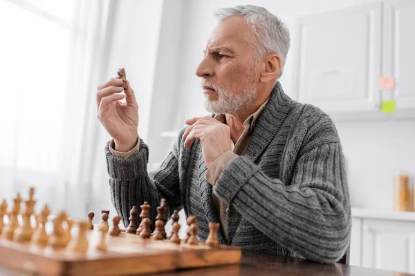 Tense man suffering from memory loss and looking at chess figure while sitting at home - foto de stock