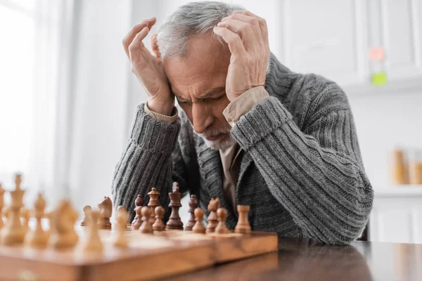 Stressed man suffering from alzheimer syndrome and sitting at chessboard with closed eyes and hands near head — Photo de stock