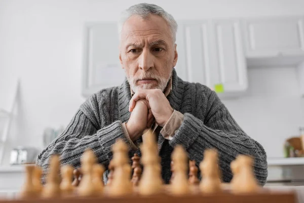 Senior man suffering from memory loss and thinking near blurred chessboard at home — Photo de stock