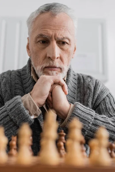 Вдумчивый пожилой человек с синдромом Альцгеймера смотрит на размытую шахматную доску дома — стоковое фото