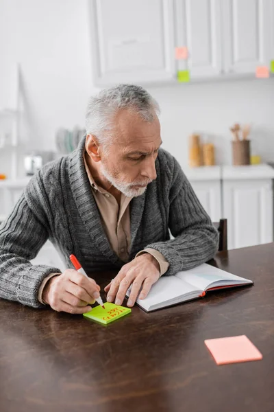Седовласый мужчина с синдромом Ацгеймера пишет номер телефона на липких нотах рядом с пустым блокнотом на столе на кухне — стоковое фото