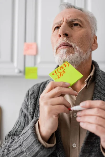 Tense man suffering from memory loss while holding sticky notes with phone number and looking away in kitchen — Stock Photo