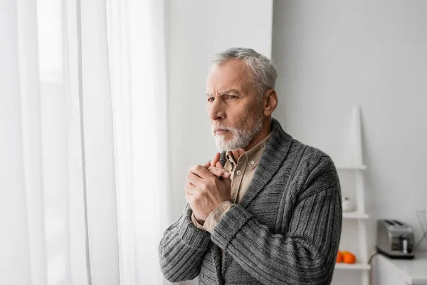 Седой мужчина с болезнью Альцгеймера стоит с сжатыми руками и смотрит в окно дома — стоковое фото