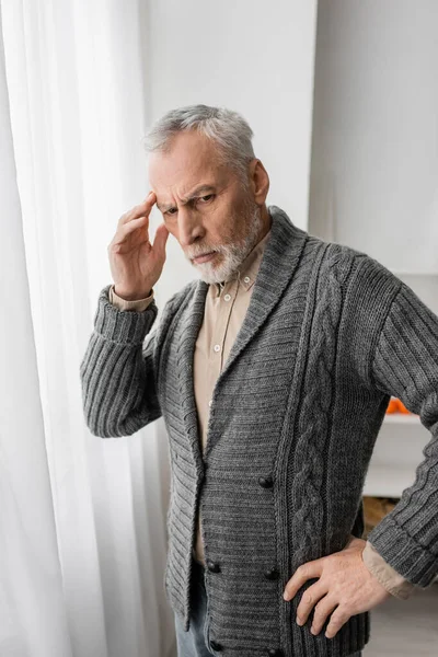 Tense man suffering from memory loss and touching forehead while standing with hand on hip at home — Stock Photo