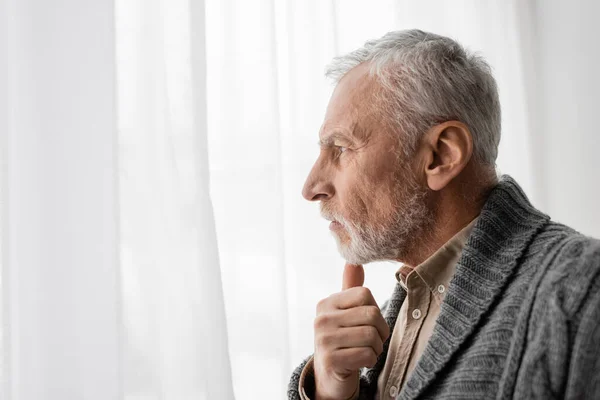 Side view of aged man suffering from memory loss and holding hand near chin while looking away at home — Stock Photo