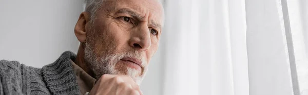Bearded senior man with alzheimer syndrome holding hand near chin and looking away at home, banner — Stock Photo