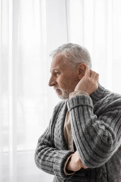 Senior and thoughtful man in knitted cardigan looking away near window while suffering from alzheimer syndrome — Stock Photo