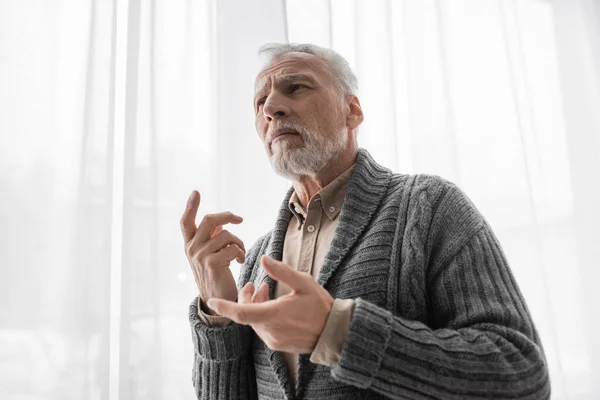 Worried senior man in knitted cardigan gesturing near window while suffering from memory loss caused by alzheimer diseased — Stock Photo