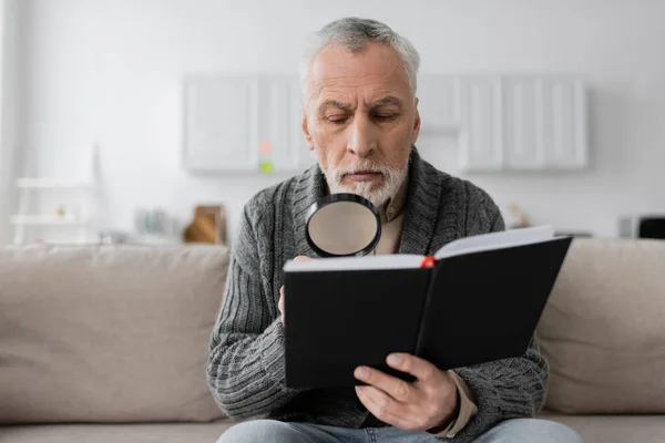 Senior man with alzheimer disease sitting on sofa and looking in notebook through magnifying glass — Photo de stock