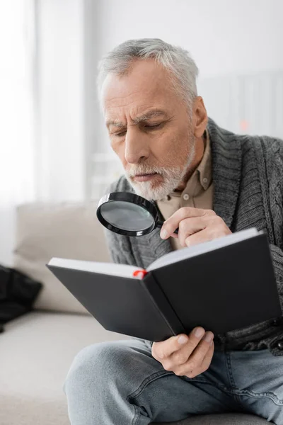 Aged man with alzheimer syndrome looking in notebook through magnifier while sitting at home — Stockfoto