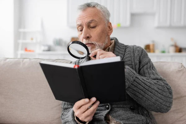 Senior man with azheimers disease looking in notebook through magnifier while sitting on couch at home — Stock Photo
