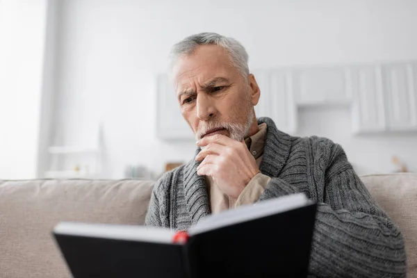 Thoughtful man suffering from memory loss and looking in notebook at home — Stock Photo