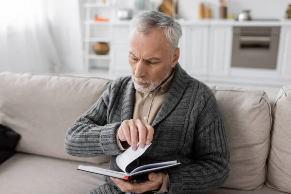 Grey haired man suffering from memory loss caused by alzheimer disease looking in notebook on couch at home — Stock Photo