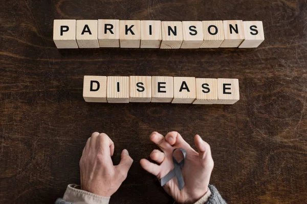 Top view of cropped man holding grey ribbon in trembling hands near blocks with parkinsons disease inscription on wooden surface — Stock Photo