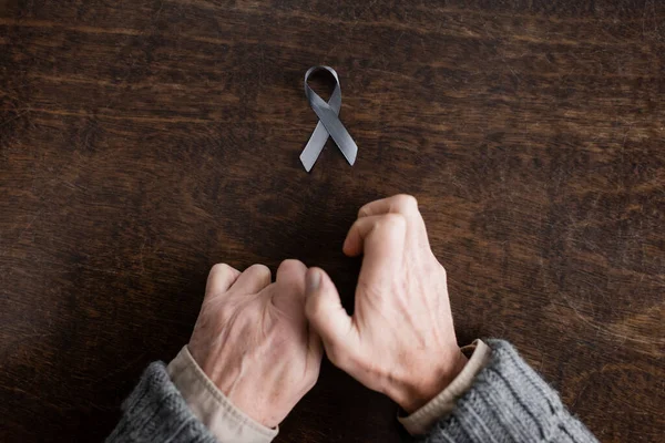 Top view of cropped man with parkinsonism and trembling hands near grey ribbon on wooden table - foto de stock