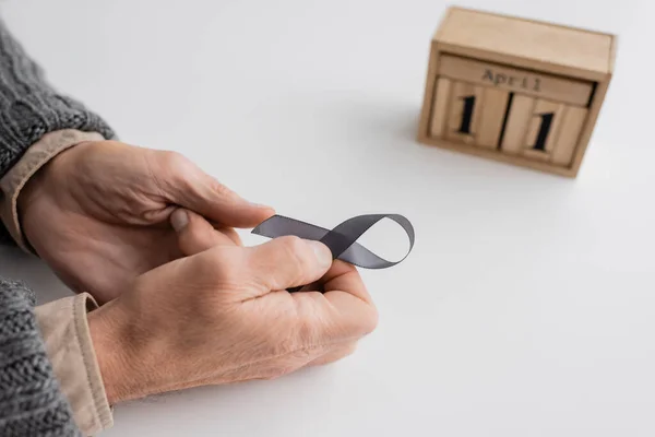 High angle view of cropped senior man with parkinsonism holding grey ribbon near wooden calendar with april 11 date on white surface — Foto stock