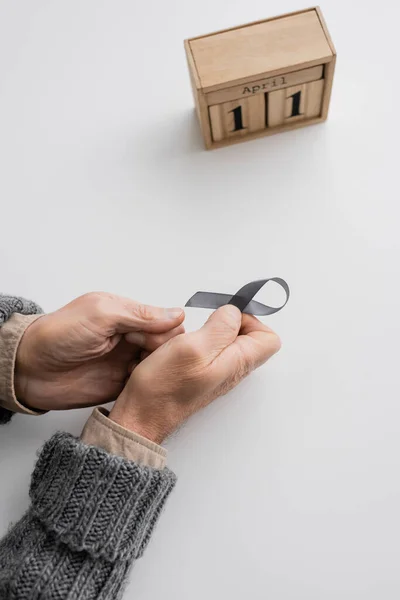 High angle view of cropped senior man with parkinson syndrome holding grey ribbon near wooden calendar with april 11 date on white surface — Fotografia de Stock