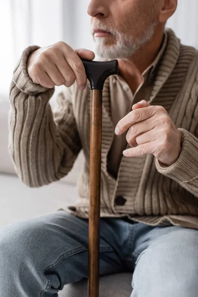 Cropped view of aged man with parkinson syndrome and hands tremor sitting with walking cane at home — Stock Photo