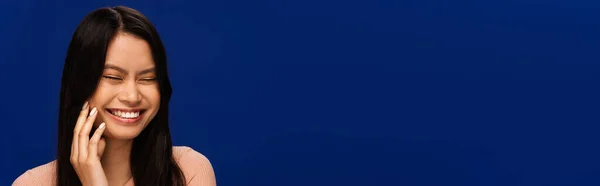 Cheerful asian woman closing eyes isolated on blue, banner — Stock Photo