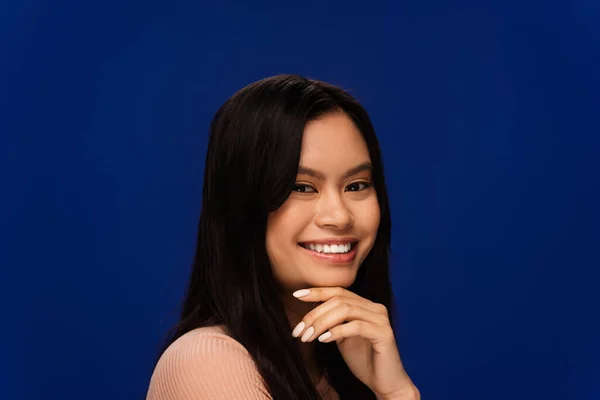 Positive asian model holding hand near chin and looking at camera isolated on blue — Stock Photo
