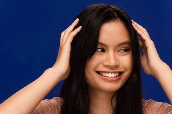 Smiling asian woman touching hair isolated on blue - foto de stock