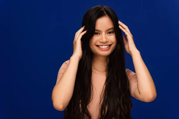 Cheerful asian woman touching long hair isolated on blue — Stock Photo