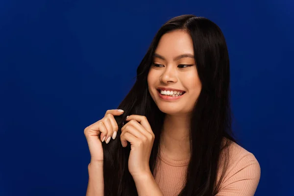 Cheerful asian woman looking at hair isolated on blue — Stockfoto