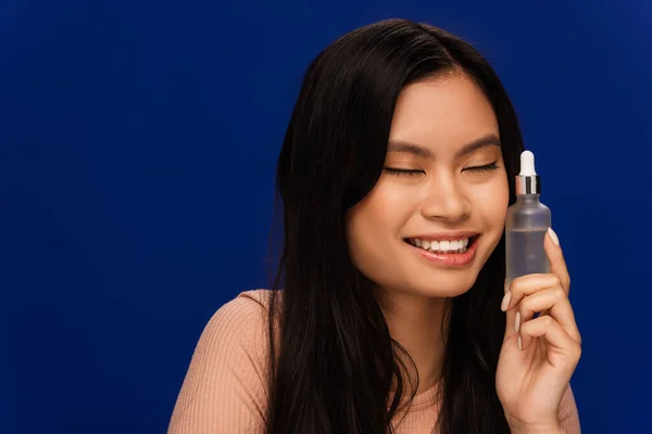 Cheerful asian woman holding bottle with cosmetic serum isolated on blue — Fotografia de Stock