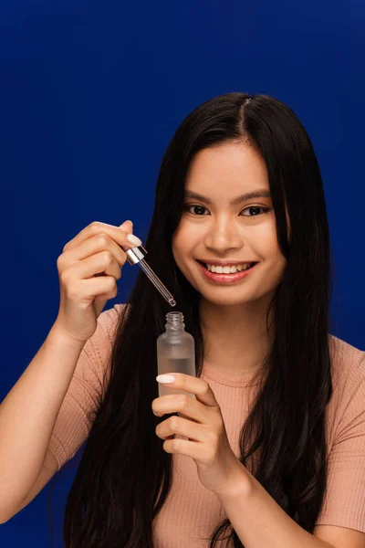 Smiling asian woman holding cosmetic serum and dropper isolated on blue — Stock Photo