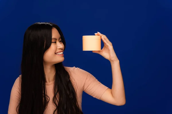 Cheerful asian woman looking at container with hair mask isolated on blue — Stock Photo