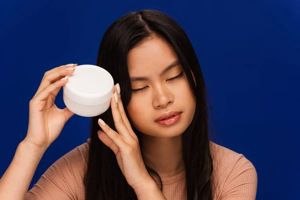 Pretty asian woman holding container with cosmetic cream isolated on blue — Photo de stock