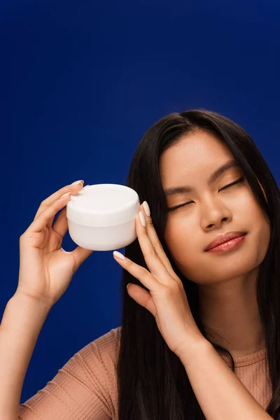 Pretty asian woman holding container with cosmetic cream isolated on blue — Stock Photo