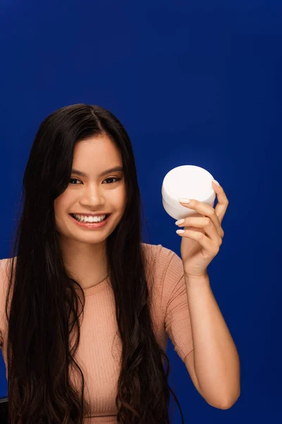 Cheerful long haired asian woman holding container with cream isolated on blue — Photo de stock