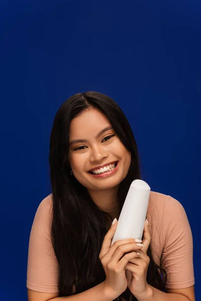 Positive asian woman in t-shirt holding hair conditioner isolated on blue - foto de stock