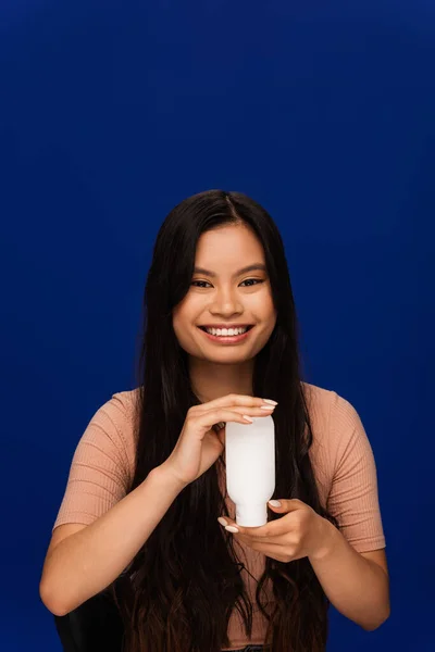 Smiling asian woman in t-shirt holding body lotion isolated on blue - foto de stock