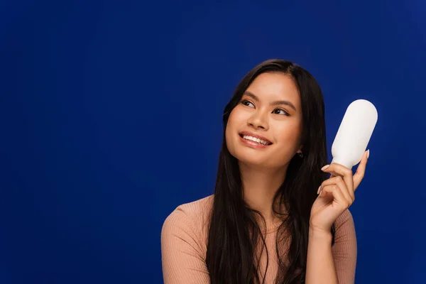 Cheerful asian model holding body lotion and looking away isolated on blue — Stock Photo