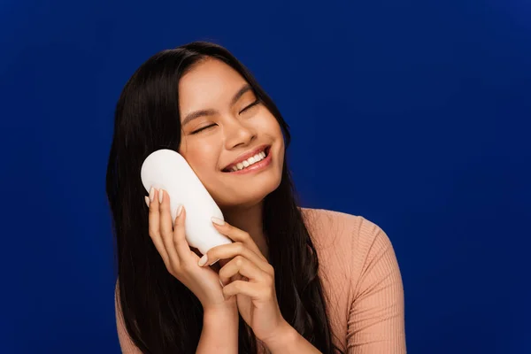 Cheerful brunette asian woman holding body lotion isolated on blue — Fotografia de Stock